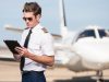 A male pilot stands outside of a corporate jet aircraft, using a modern digital tablet to update his flight plan and preflight checklist.